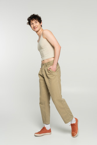 carefree bigender person in tank top and sneakers posing with hand in pocket of beige pants on grey.jpg