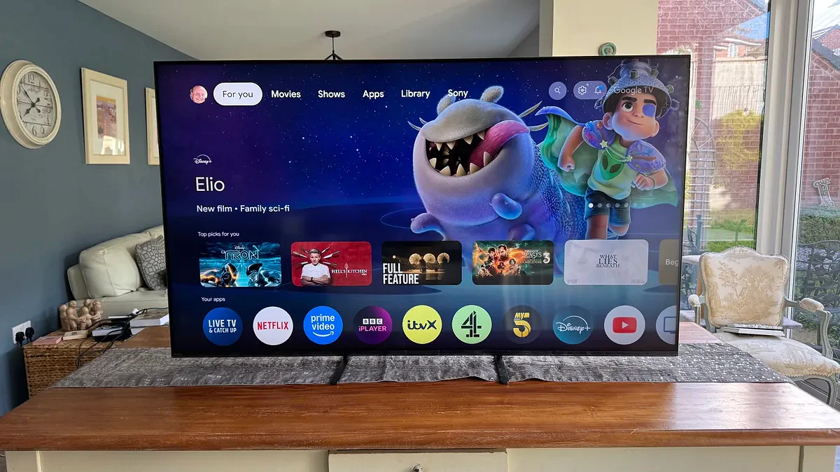 The Sony 65 Bravia 5 TV showing the home screen from a distance, positioned on a wooden cabinet