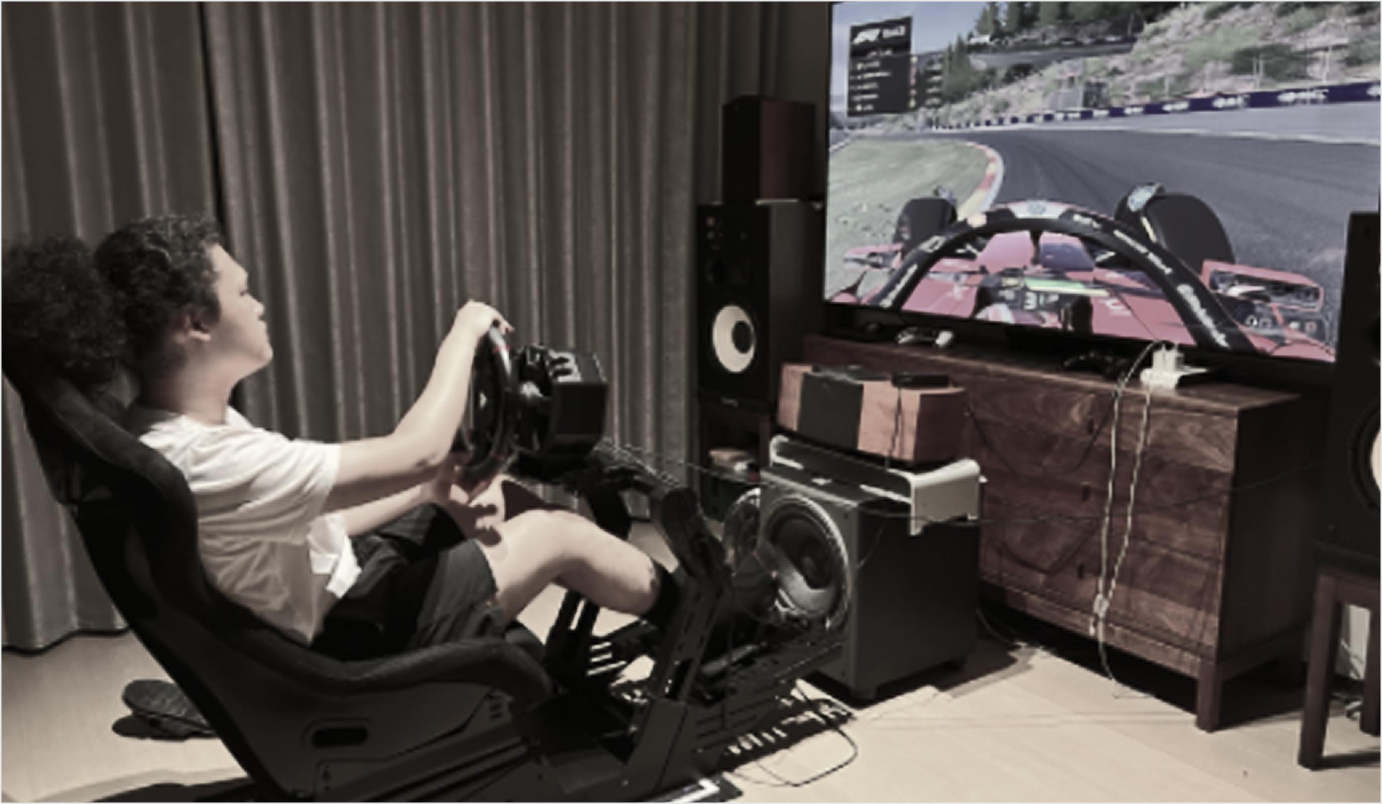 Side view of a driver in a bucket seat with wheel and pedals, watching an F1 game