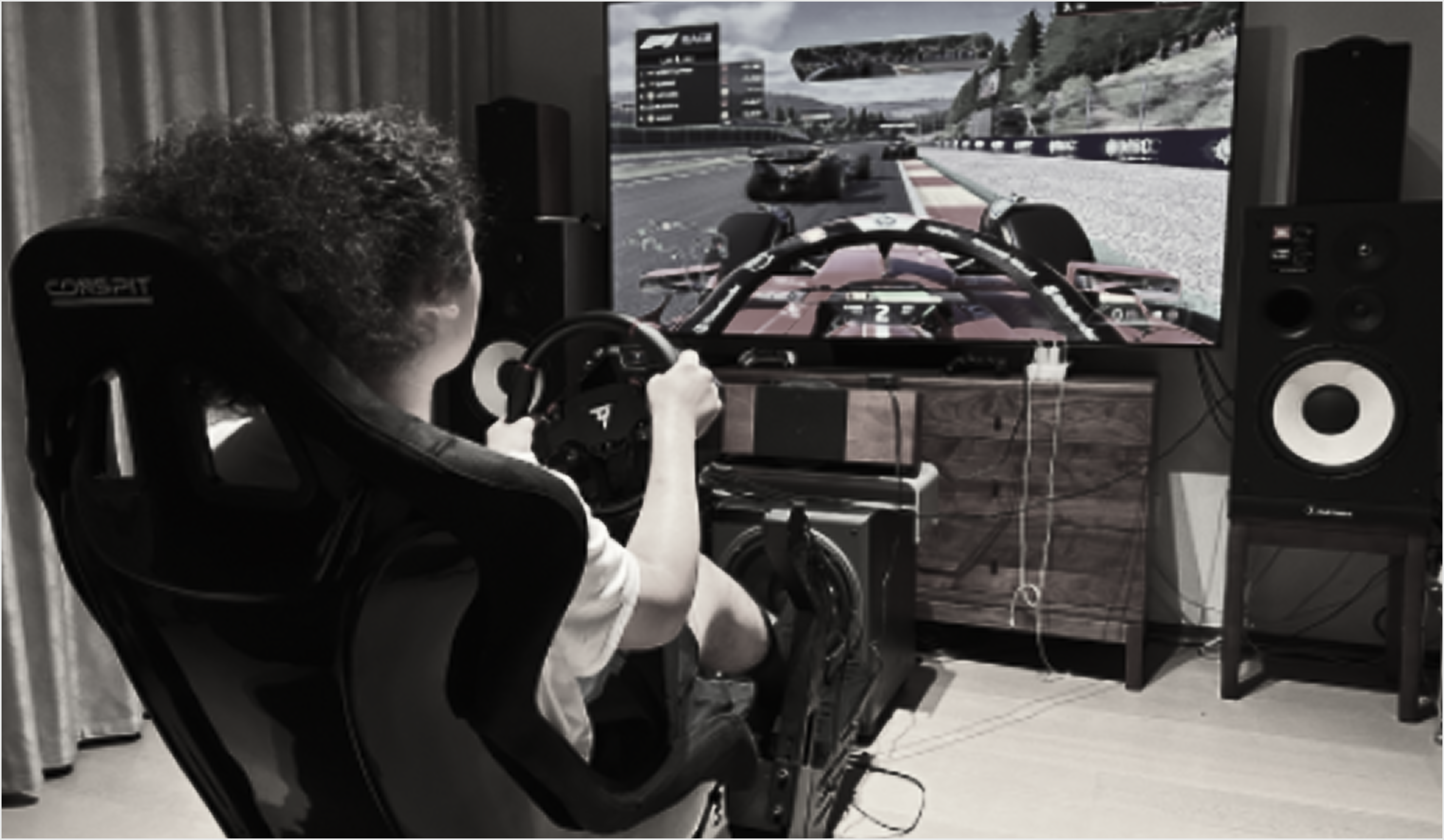 Rear view of a driver in a home racing simulator, holding the wheel while an F1 cockpit is on the TV