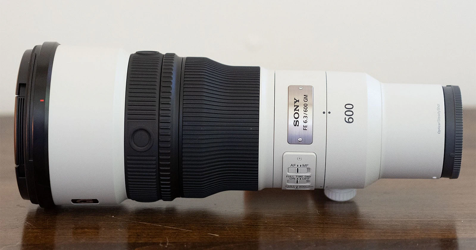 A white Sony FE 6.3/600 GM telephoto camera lens sits horizontally on a dark wooden surface against a plain white background.