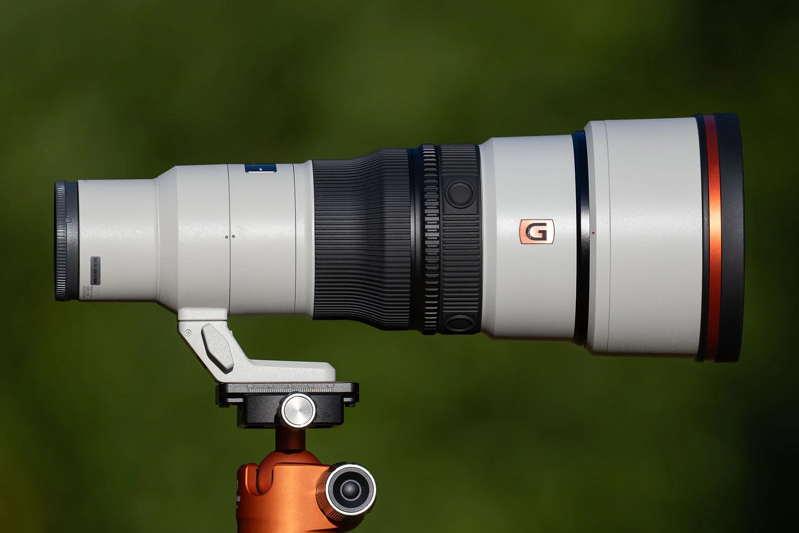 A long, white telephoto camera lens with a black focus ring and a prominent orange "G" logo, mounted on an orange tripod head against a blurred green background.