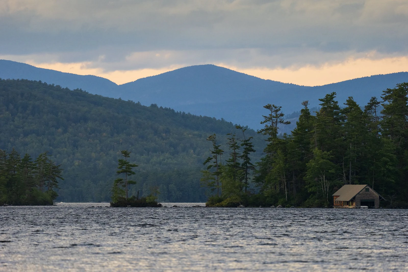 A boathouse sits on the shore of a lake with small islands and forested mountains in the background under a cloudy sky.