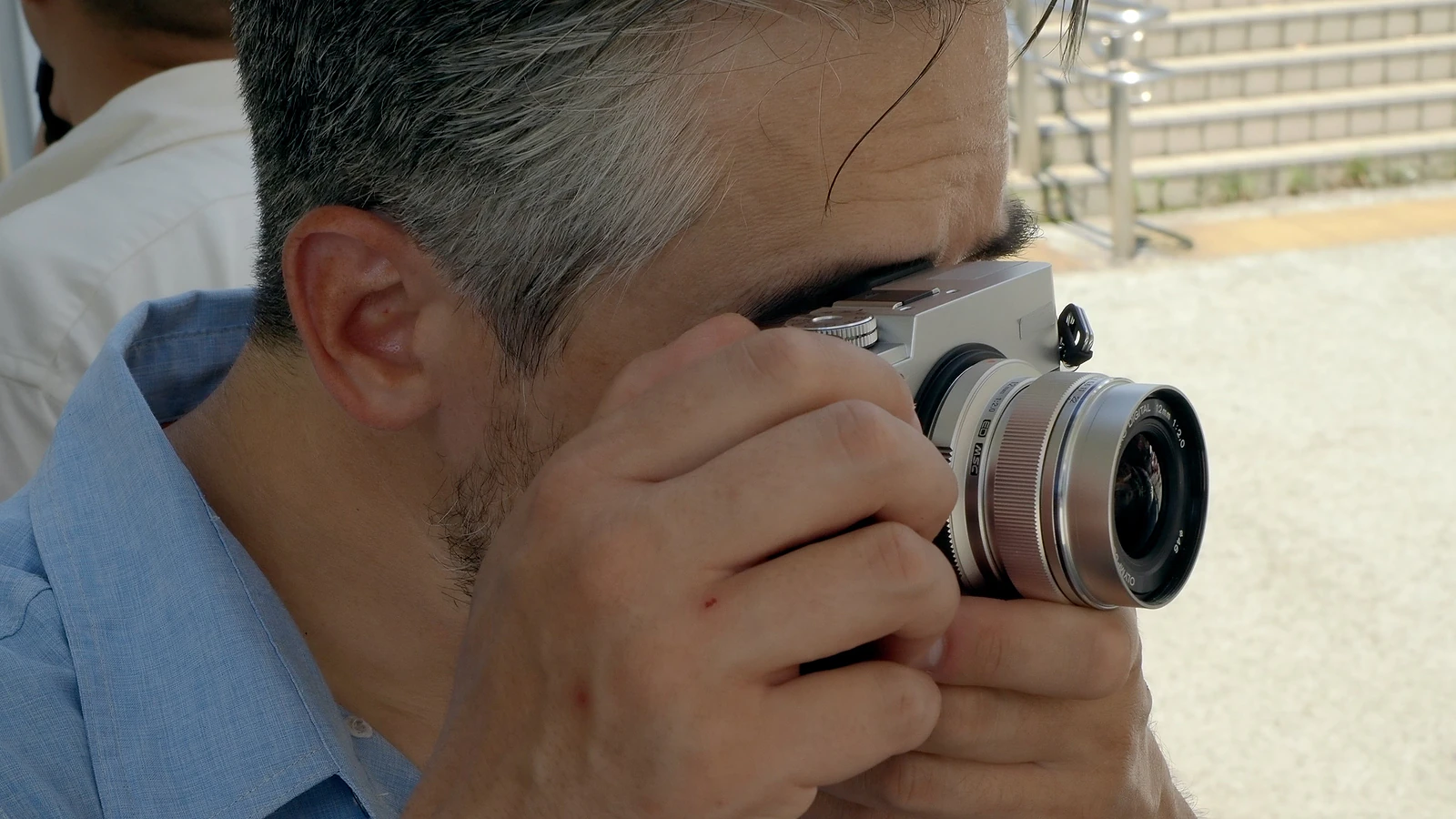 A man with graying hair holds a silver digital camera to his eye to take a photograph.