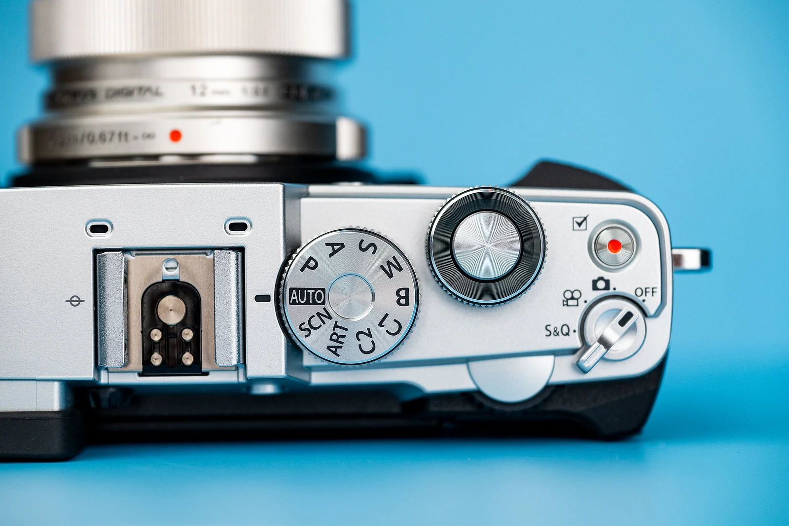 A top-down view of a silver digital camera body with a lens, featuring a mode dial, shutter button, and power switch against a blue background.