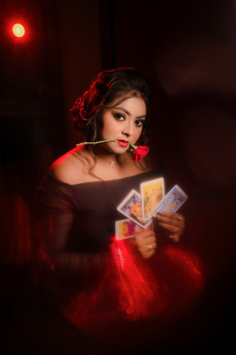 Priyadarsini Mallikk holding a fanned four tarot cards, a red rose held in her mouth, lit against a dark background