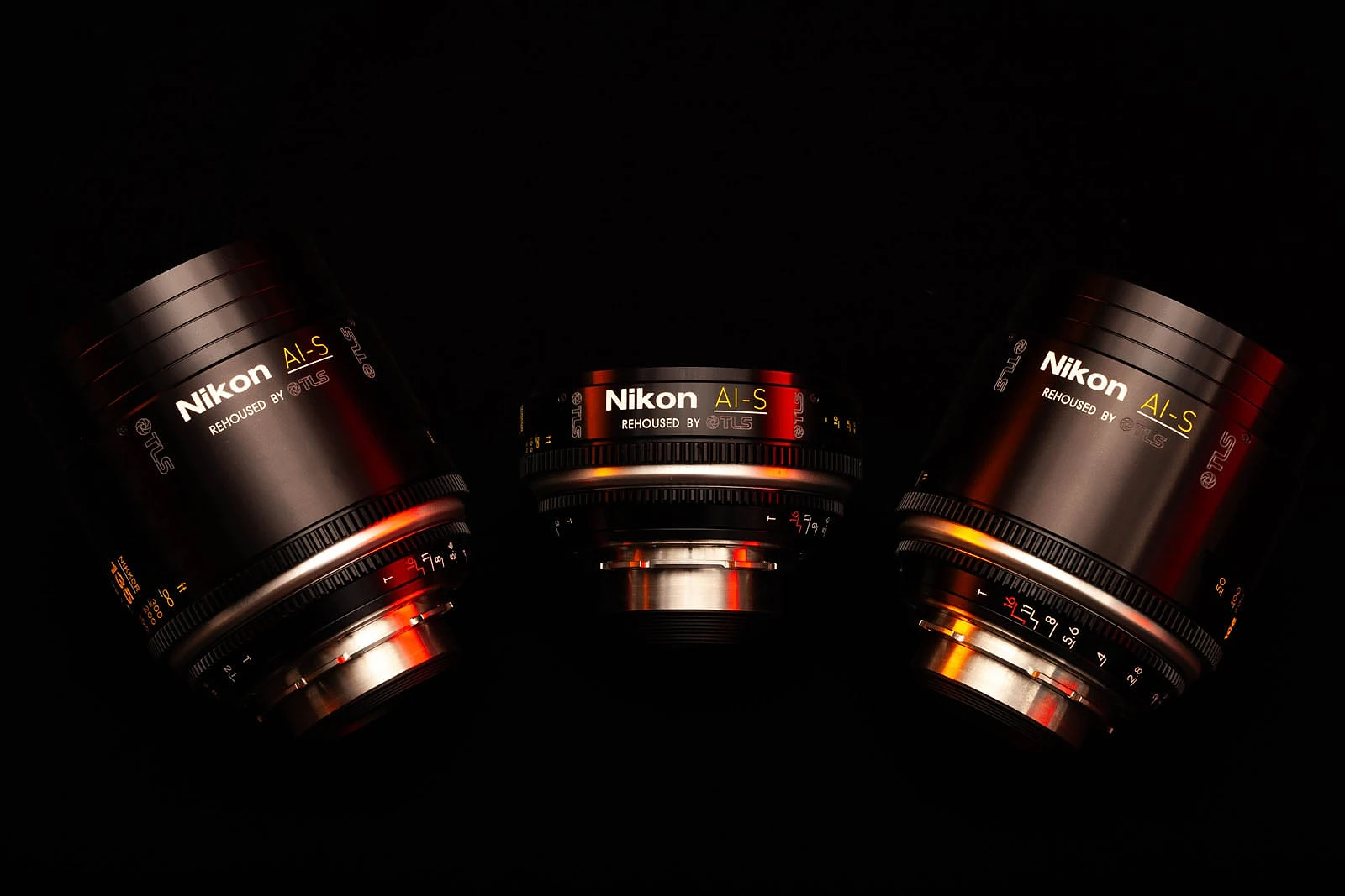 Three Nikon AI-S lenses rehoused by TLS are arranged against a black background with red and orange lighting accents.