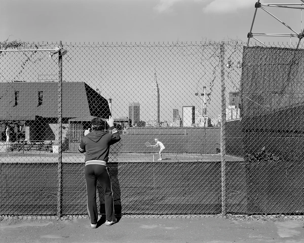 A man in a tracksuit grips a chain-link fence to watch a tennis match, with the Manhattan skyline visible beyond the court.
