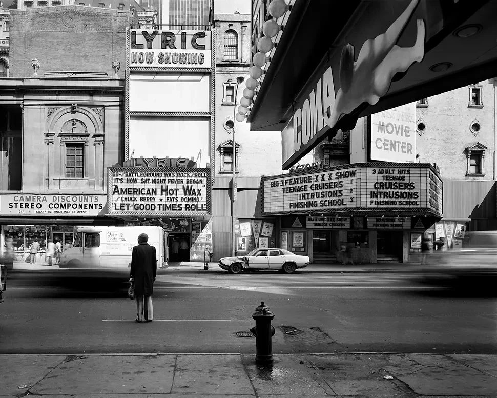 A man in a long coat stands alone at a street corner beside a fire hydrant, facing a row of cinema marquees advertising old films.