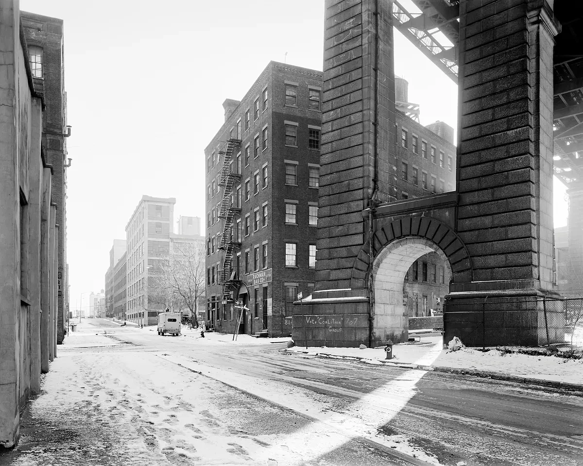 A snow-covered city street runs beneath a stone archway and elevated railway trestle, with a small van parked in the distance.
