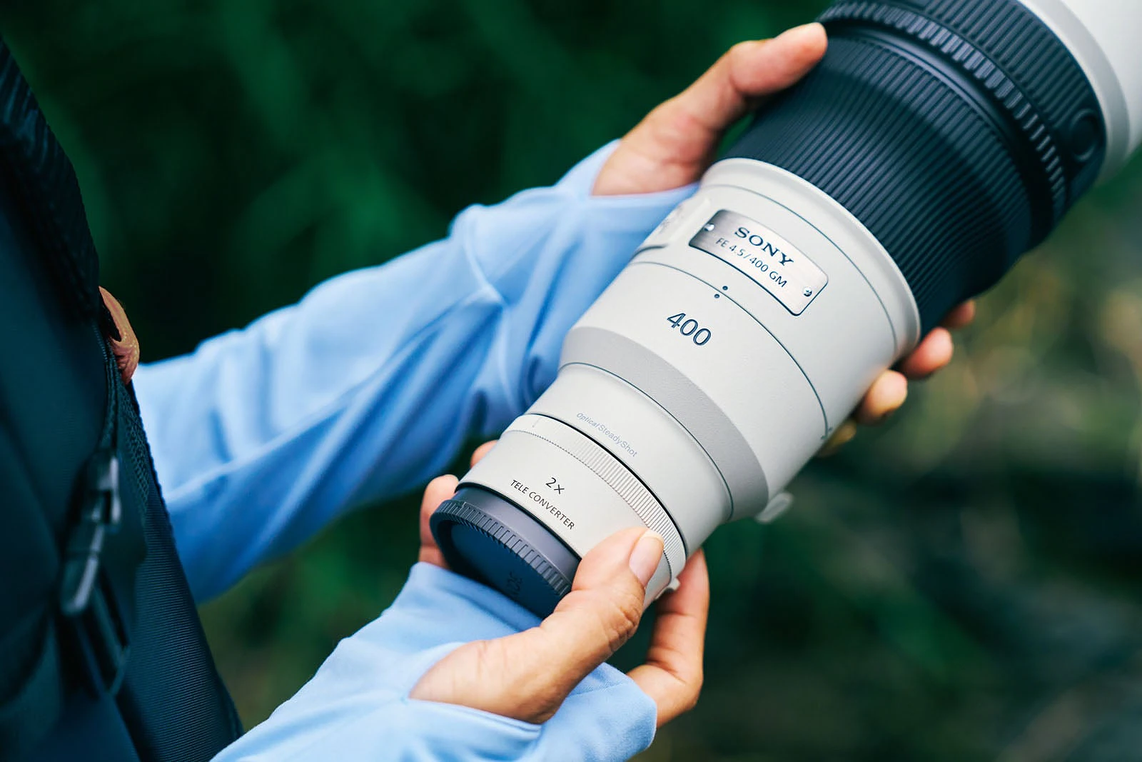 A person wearing a light blue long-sleeved shirt holds a white Sony FE 4.5/400 GM lens with a 2x teleconverter attached.