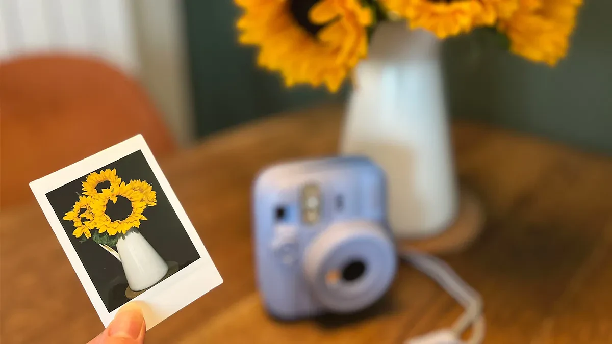 The Frost Blue Instax Mini 13 on a table next to a vase of sunflowers with a printed picture of the sunflowers being held in focus