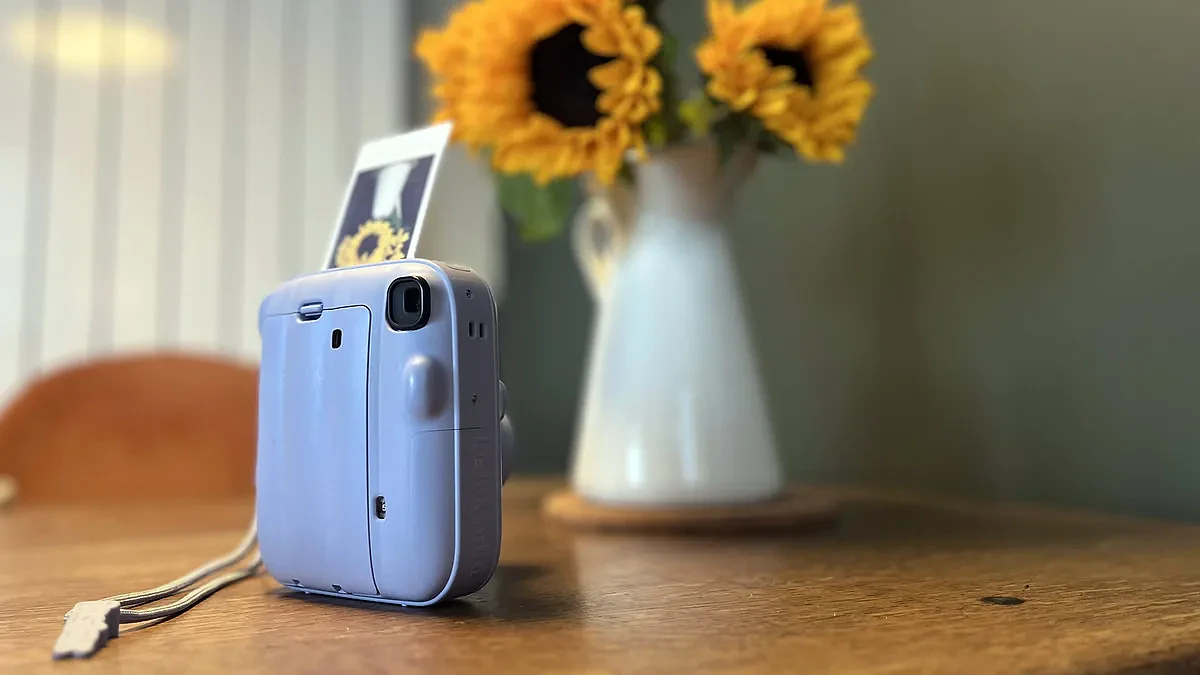 The Frost Blue Instax Mini 13 on a table with a print sticking out of the top of it next to a vase of sunflowers