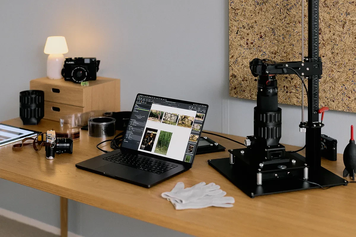 Workspace with a laptop displaying photo editing software, cameras, camera repair tools, and white gloves on a wooden desk