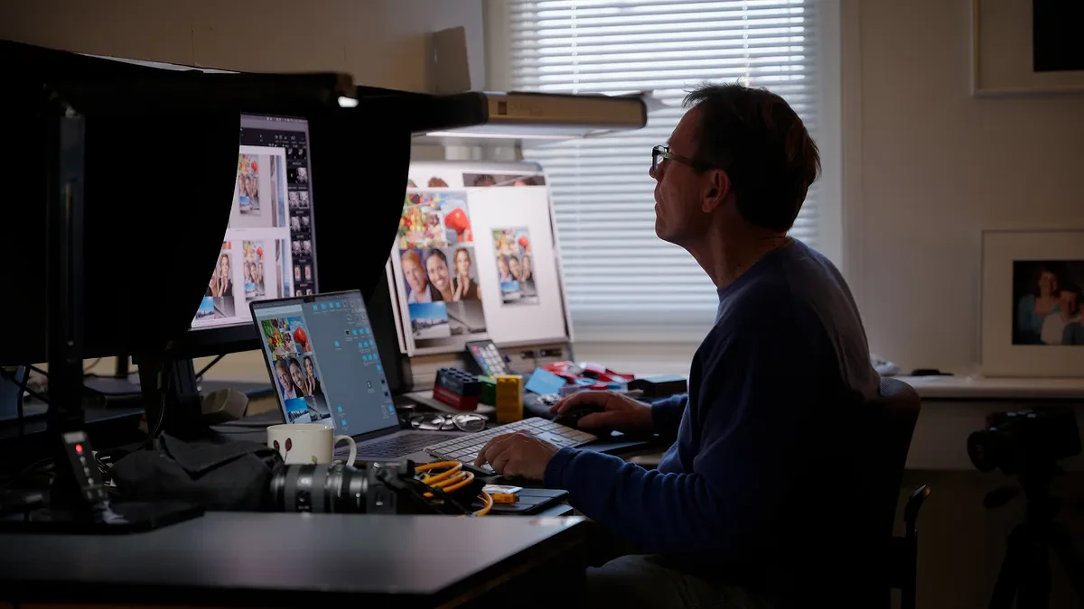 Man working on photo editing at a dual-monitor desk setup in a dimly lit room with photography equipment nearby