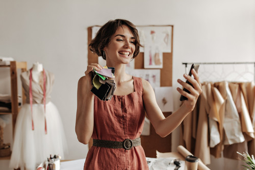 Joyful young brunette short-haired woman in linen red dress smiles sincerely, holds pieces of textil.jpg