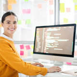 Pretty young webdesigner in casualwear looking at you by her workplace while working over new projec