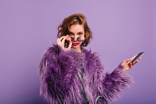 Interested girl in stylish purple fur coat looking to camera holding sparkle sunglasses. Indor portr.jpg