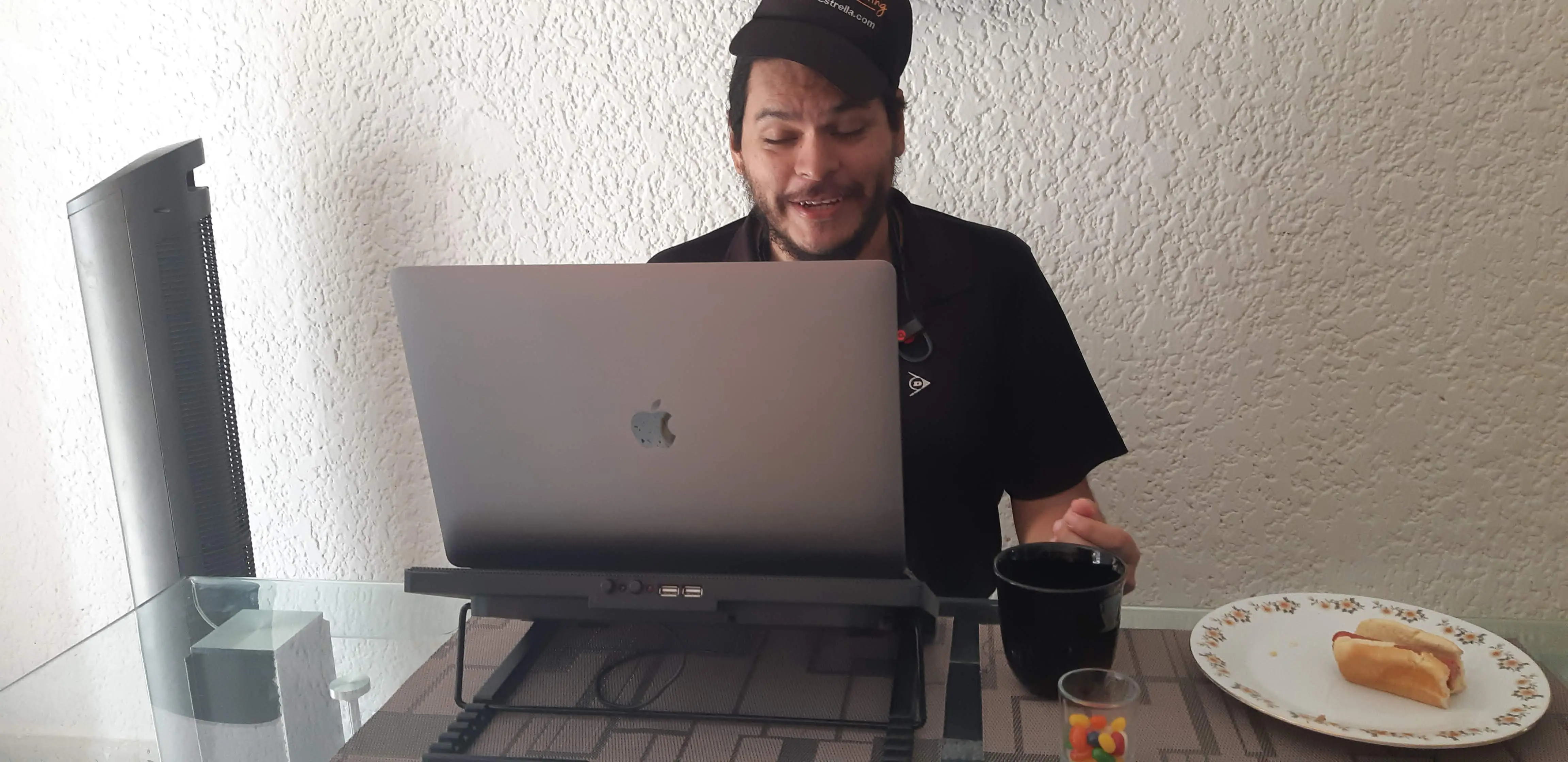 Trabajando frente a la laptop con la gorra mal puesta, los ojos cerrados; en la mesa: café, jelly beans y medio hot-dog